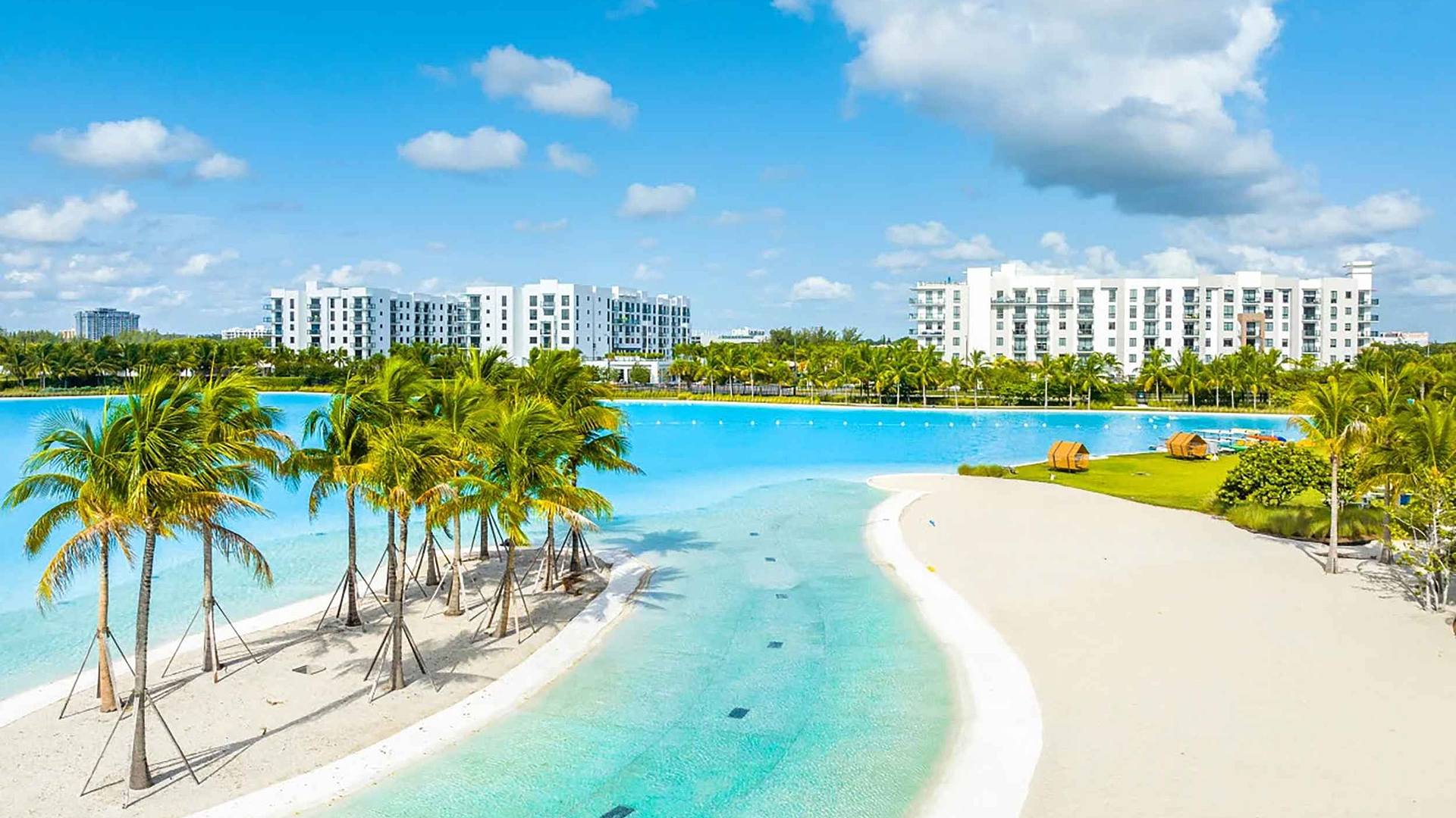 Laguna by Sole Mia Rentals A serene tropical beach with palm trees, clear blue water, and modern buildings in the background under a sunny sky.